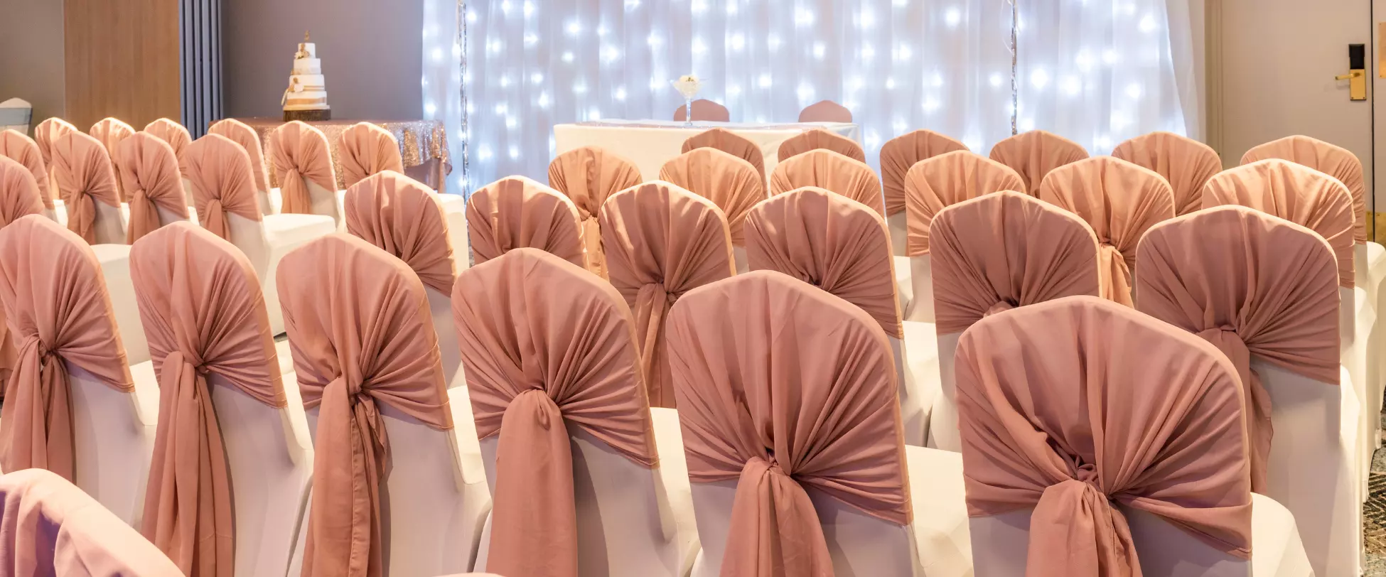 Rows of chairs set up for a wedding ceremony. The chairs are covered in white with dusty pink bows. There is a table at the front with two chair for the couple..