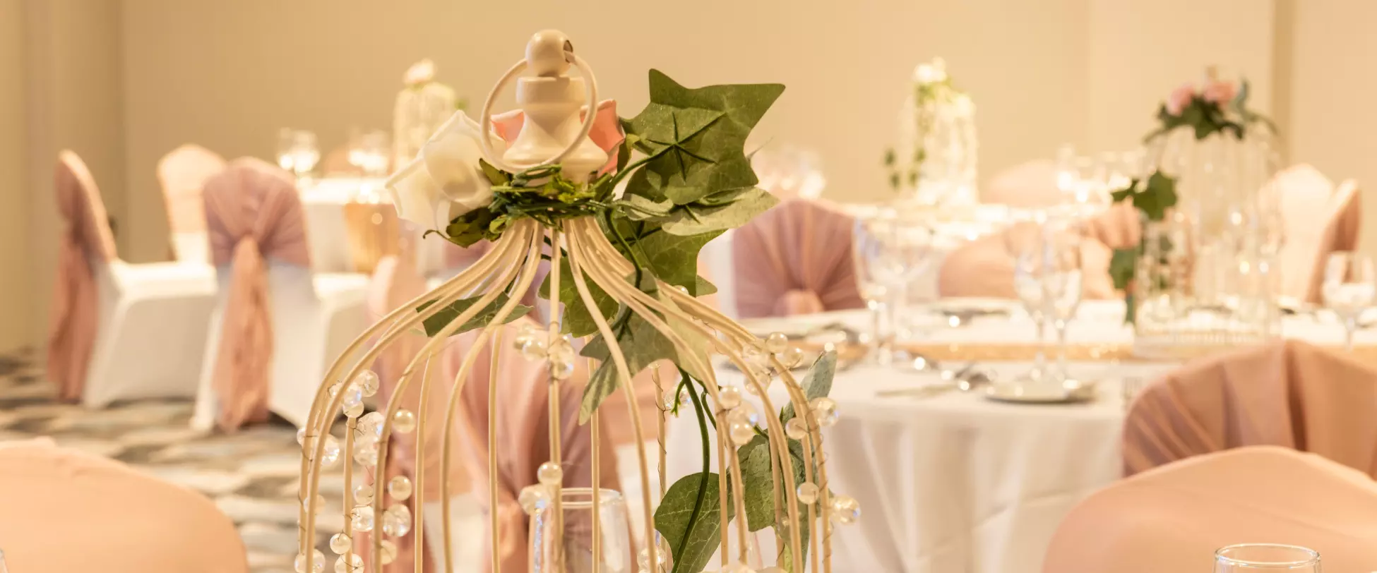 A gold bird cage sits in the middle of a circular table set up for a wedding. In the birdcage is a floral arrangement with pink bows..