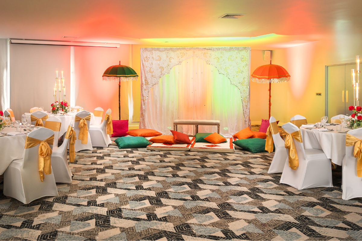 Circular tables dressed with white linens and gold sashes surround a central area that has green orange and pink cushions, a two seater bench, and colourful umbrellas..