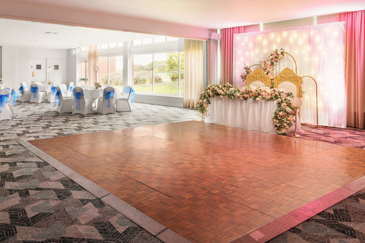 A large room is dressed for a wedding. A table to the right has two large gold and cream chairs and has white and pink flowers draping around. On the left are circular tables set up for a wedding meal and in the corner a table is set up for a buffet..